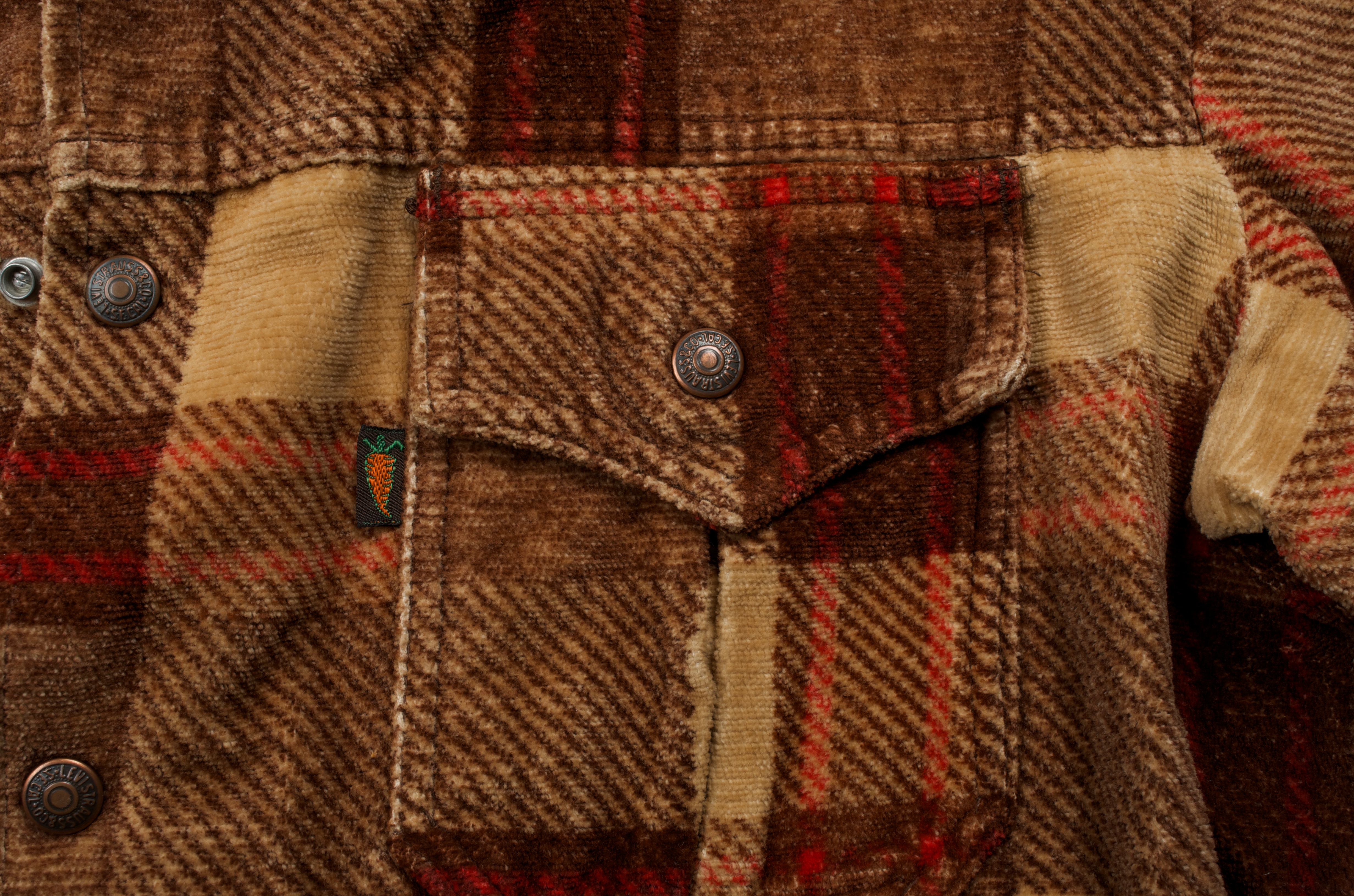 A close up of the front pocket on a Levi's® flannel button down in browns and reds. There is a Levi's® tab on the pocket with an orange carrot stitched in thread. 