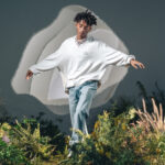 Jaden Smith poses with his eyes closed and armed stretched out wearing a stack of necklaces, a white Levi's® pullover reading "There's No Place Like Earth" and blue denim Levi's® jeans. He walks through a field of shrubbery and flowers in front of a dark sky background. An abstract amorphous white shape is behind his torso.