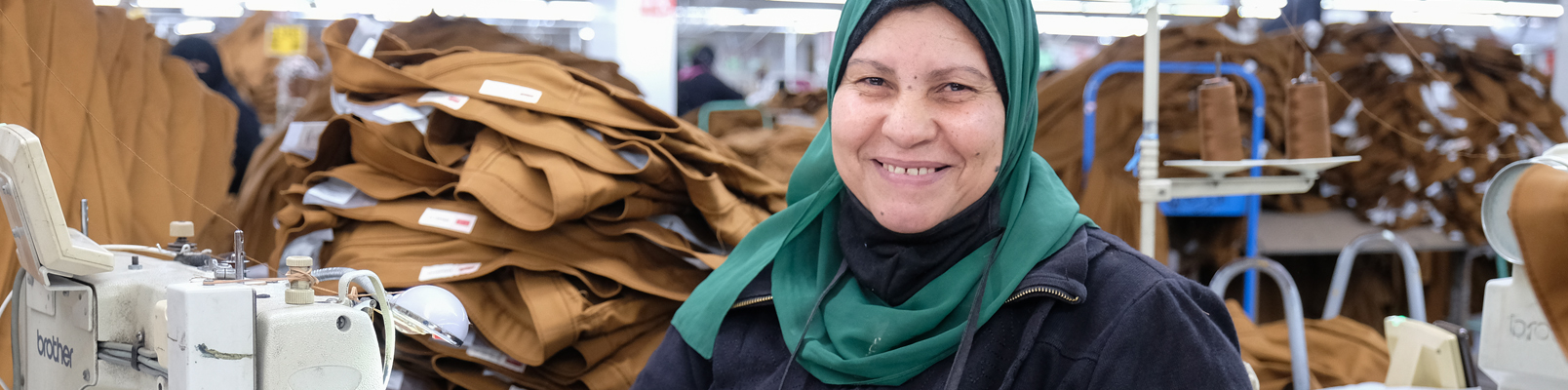 A person in a retail factory setting next to a pile of stacked pants smiles at the camera wearing a green headscarf