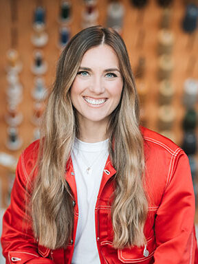 A headshot of LS&Co. Chief Merchandising Officer Dawn Vitale. She wears a red jacket over a white shirt with blue denim Levi's® jeans. An out-of-focus wall of spools of threads is behind her.