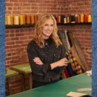 LS&Co. CEO Michelle Gass smiles with her arms crossed. She is wearing a black denim Levi's® jacket. There is a brick wall with spools of thread behind her. Her photo is overlayed on top of a blue denim texture background.