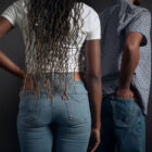 The torso and top half of legs of a person with long brown braids faces away from the camera. Their left hand is in the back left pocket of their blue denim Levi's® jeans and they wear a white T-shirt. Slightly behind and to the right of them, another person's torso faces away from teh camera. Their right hand is in the back pocket of their Levi's® denim jeans and they are wearin ga blue and white floral pattern shirt.