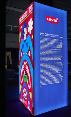 A large blue illuminated rectangular block in the new Levi's® exhibition, Icons, Innovations & Firsts — Stories of Heritage and Progress From the Levi’s® Archives. On the left of the block is the colorful poster art for the exhibition, the right side features the Levi's® logo at the top followed by a large text description of the exhibit.
