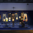 A section of the new Levi's® exhibition, Icons, Innovations & Firsts — Stories of Heritage and Progress From the Levi’s® Archives. Various Levi's® pieces are displayed in a dark room with a blue background. Description plaques are placed in front of each item.