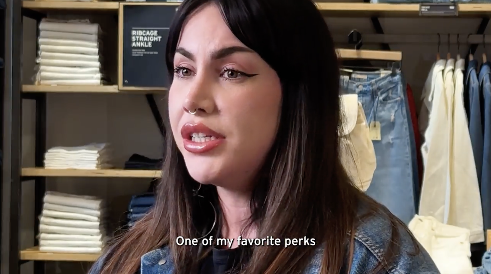 A close up of Kaitlyn Fernandez, assistant store manager for the Levi's® brand, mid speech. She is inside a Levi's® store in front of a shelf of Levi's® jeans and a text caption reads 
