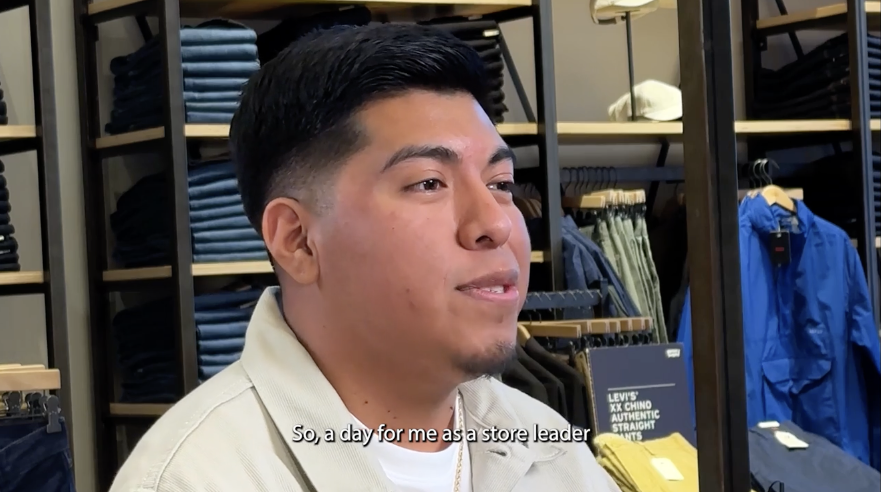 A close up of Omar Martinez, assistant manager for the Levi's® brand. Behind him, a shelf of folded Levi's® jeans. He is wearing a beige collared shirt over a white T-shirt and a caption reads 