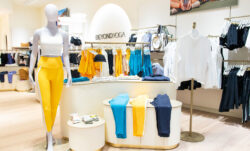 The floor of Beyond Yoga® Bellevue. A mannequin wears a white Beyond Yoga® tank and yellow Beyond Yoga® leggings. Blue, yellow and black leggings are folded on a center display. Assorted products are seen hanging in the background.