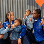 A family of five sits on a sidewalk. A mother with long hair holds two young children, a father holds an orange balloon and a blue balloon in one hand and a child who is eating an ice cream cone. The parents are smiling at each other and wearing matching blue satin jackets with New York mets logos from the Levi's® x Starter collaboration