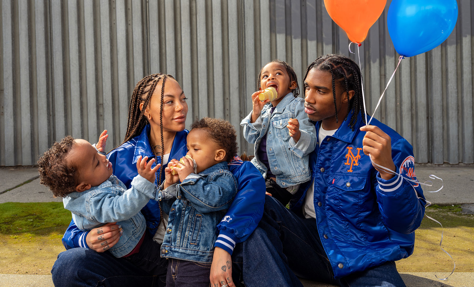 A family of five sits on a sidewalk. A mother with long hair holds two young children, a father holds an orange balloon and a blue balloon in one hand and a child who is eating an ice cream cone. The parents are smiling at each other and wearing matching blue satin jackets with New York mets logos from the Levi's® x Starter collaboration