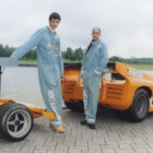 Two people pose in the Levi's® x McLaren Racing racing suits and each lean on a bright orange sports car.