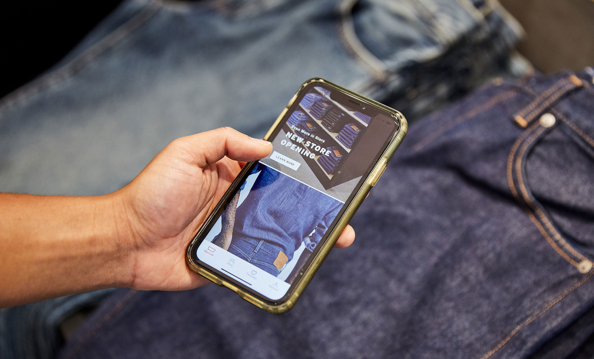 A hand holds an iPhone over two pairs of laid out jeans. The Levi's® app is opened on the phone screen, featuring a shelf of stacked jeans and the text "Even More in Store, New Store Opening, learn more."