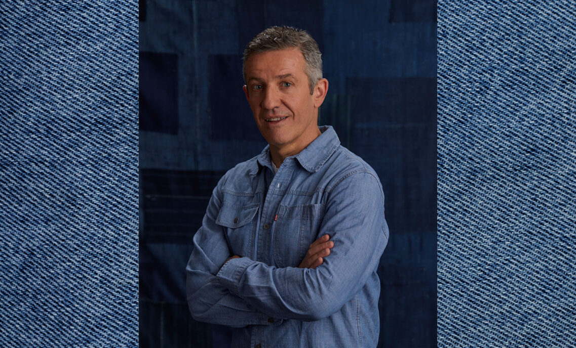 LS&Co. Chief Commercial Officer Gianluca Flore smiles and poses with his arms crossed in front of a blue background. The photo is imposed on top of a blue denim background.