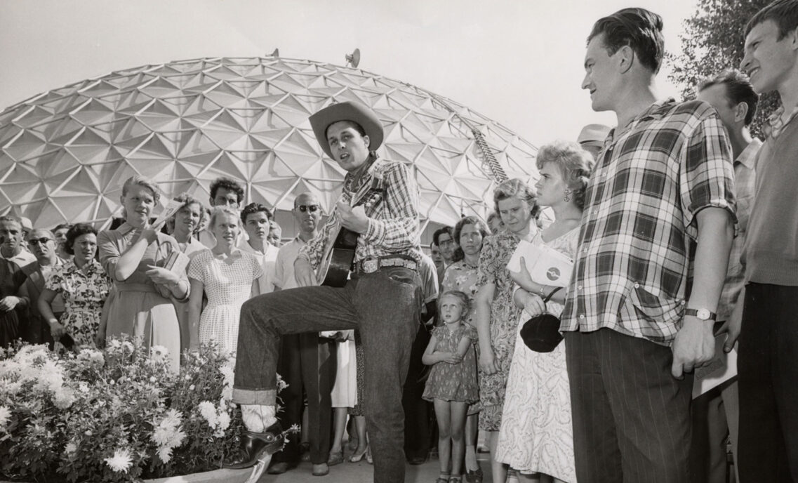 A black and white image of a man wearing a cowboy hat and Levi's® jeans playing the guitar as onlookers watch the performance