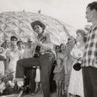 A black and white image of a man wearing a cowboy hat and Levi's® jeans playing the guitar as onlookers watch the performance