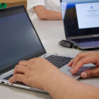 A pair of hands type on a laptop keyboard. Another laptop can be seen in the background.