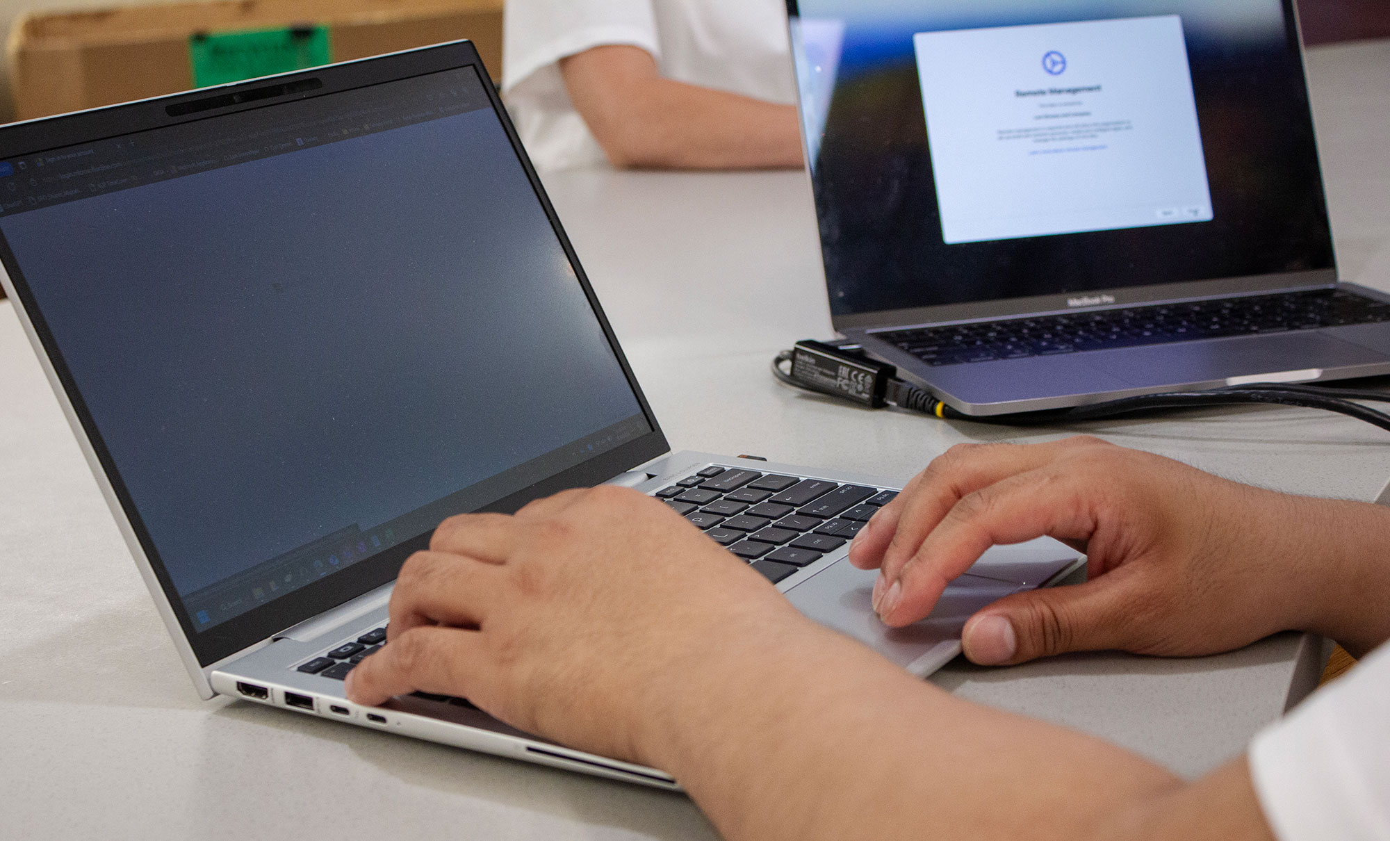 A pair of hands type on a laptop keyboard. Another laptop can be seen in the background.