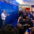 A person stands against a blue background that reads "Denim Authority" and speaks into a microphone facing a media crowd of people with phones and cameras.