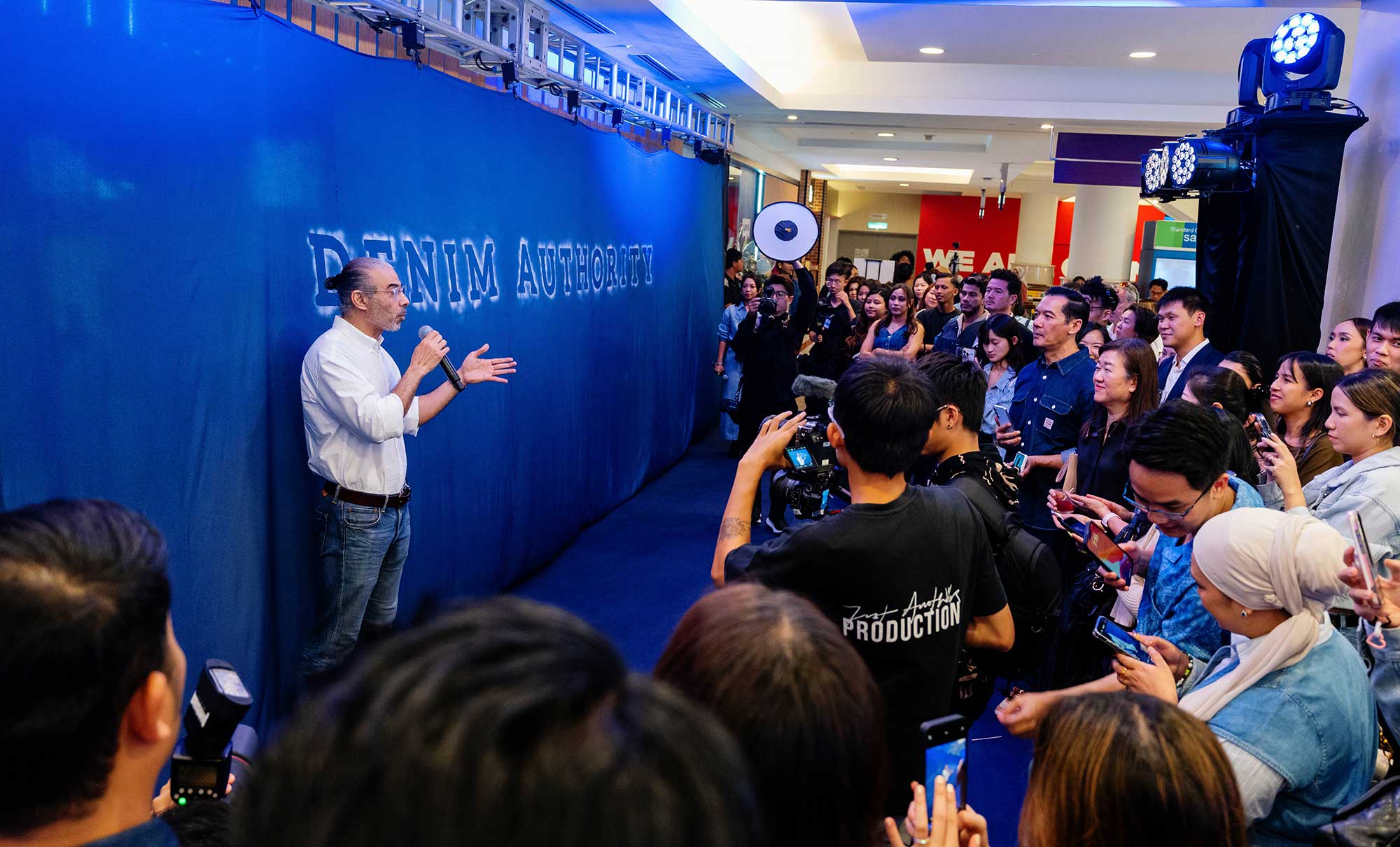 A person stands against a blue background that reads "Denim Authority" and speaks into a microphone facing a media crowd of people with phones and cameras.