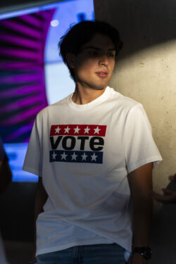 A person wearing a white T-shirt reading "VOTE" leans against a cement pillar and looks to the right. A shadow slightly covers their face and their hands are behind their back.