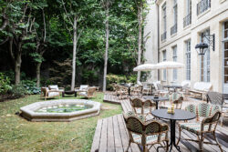 The back patio of the Levi's® Haus of Strauss Paris, featuring Parisian cafe tables and chairs with umbrellas, a fountain pond and tree-lined lawn