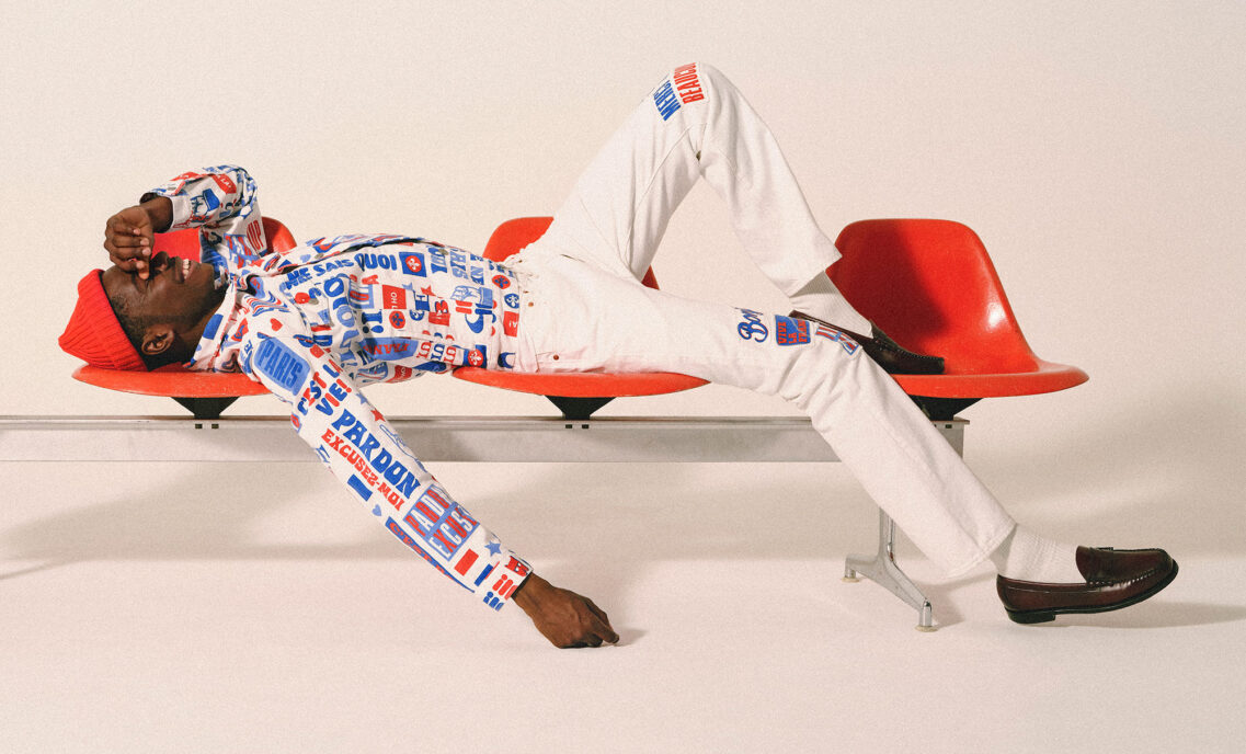 A model lies across a red bench and wears white pants and a red, white and blue long sleeve shirt with Paris motifs and text from the Levi's® x Paris collection.