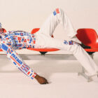 A model lies across a red bench and wears white pants and a red, white and blue long sleeve shirt with Paris motifs and text from the Levi's® x Paris collection.