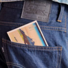 Close up of a person's back pocket. They wear dark wash denim jeans from the Levi's® Blue Tab collection, featuring a LS&Co. leather back patch. A postcard peeks out of the pocket.