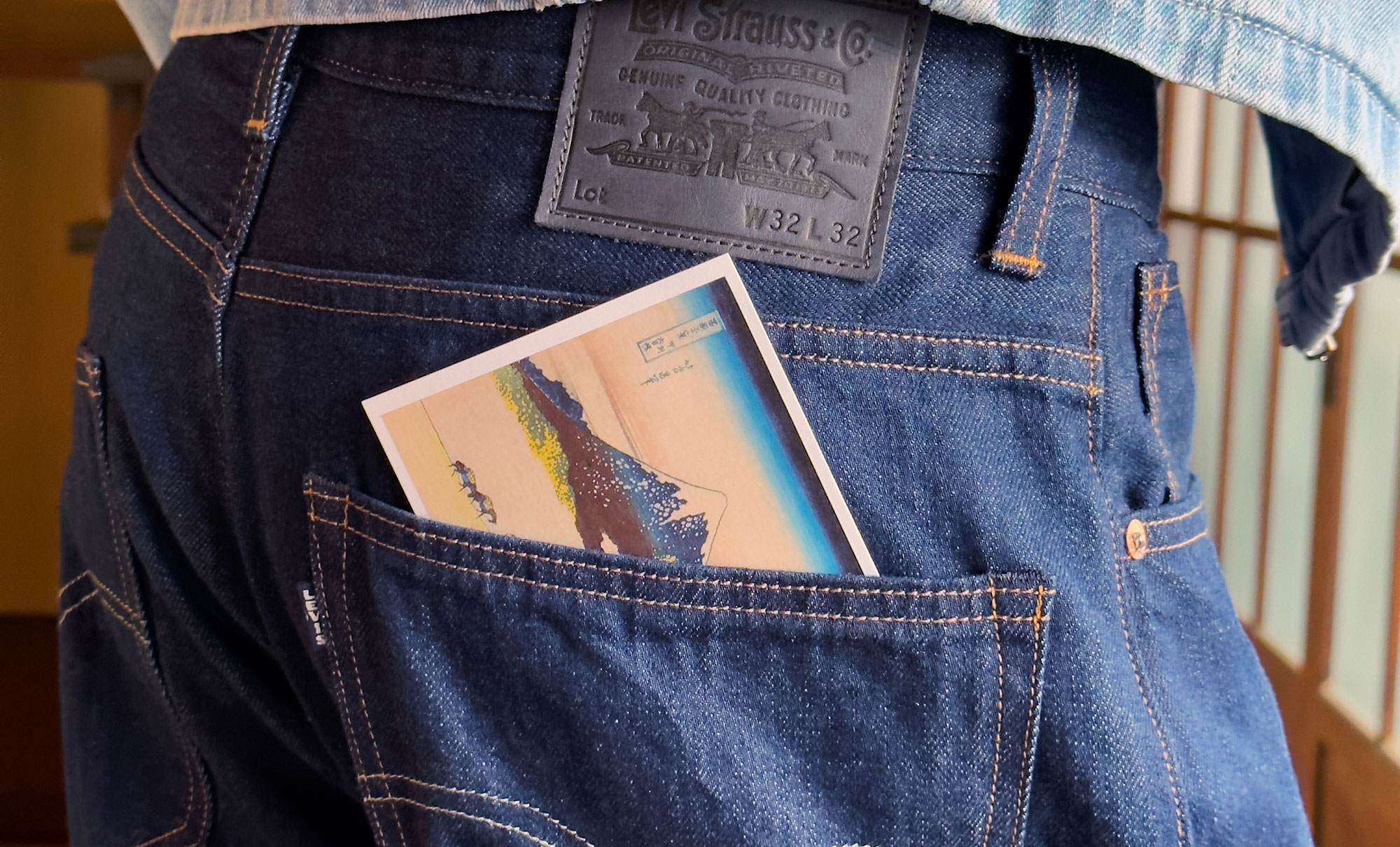 Close up of a person's back pocket. They wear dark wash denim jeans from the Levi's® Blue Tab collection, featuring a LS&Co. leather back patch. A postcard peeks out of the pocket.