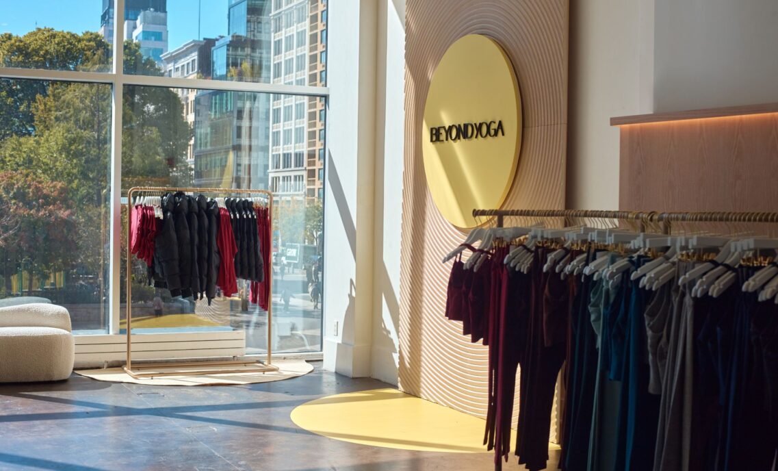 Inside a Beyond Yoga store. A rack of Beyond Yoga product sits in front of a window. A Beyond Yoga logo in a yellow circle is on the wall, next to another rack of Beyond Yoga product