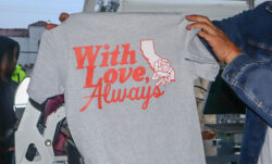 Hands hold up a grey t-shirt with red text reading "With Love, Always" next to a graphic of the state of California overlaid with a rose