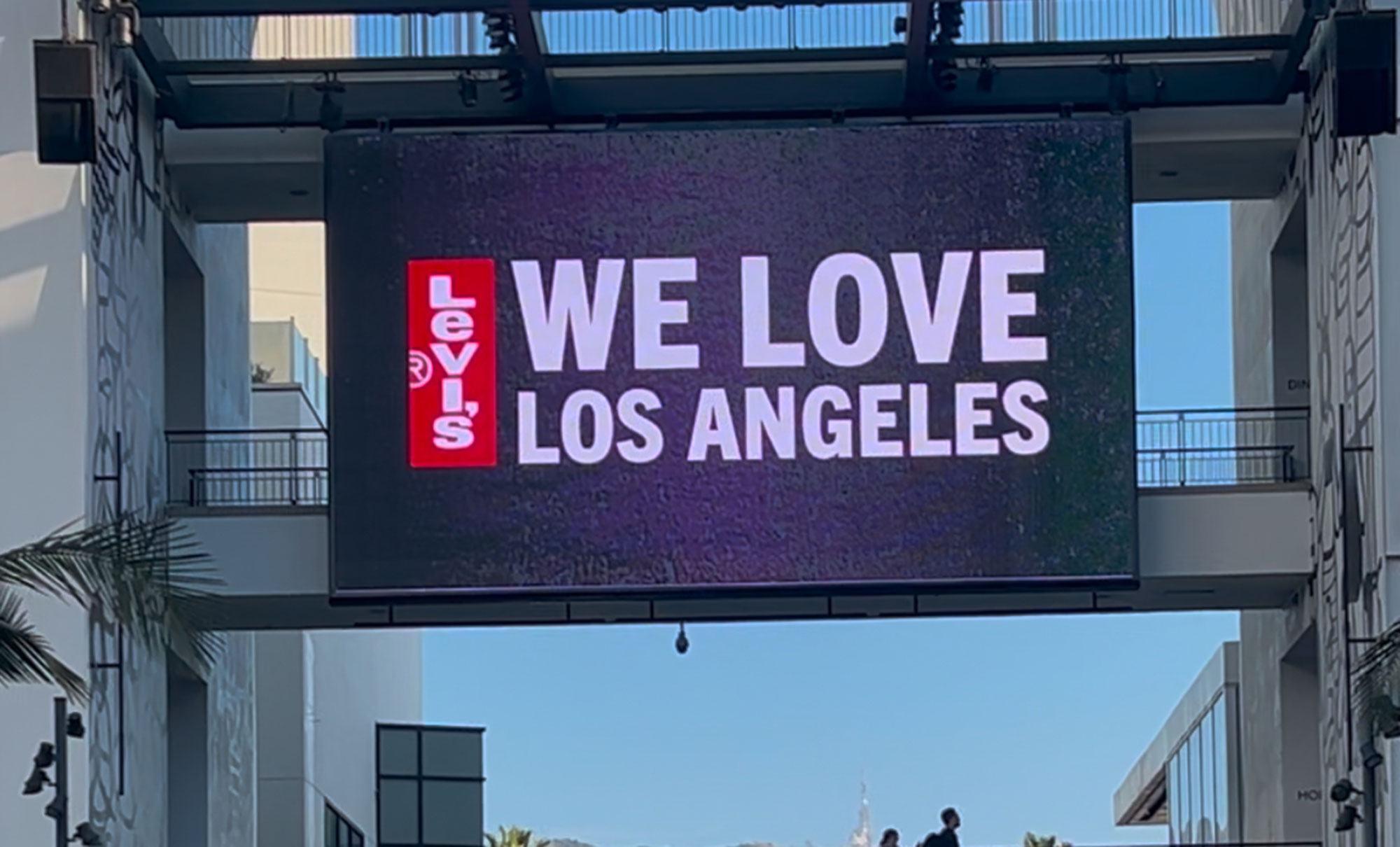 A digital sign with a Levi's® red tab logo and the text "We Love Los Angeles" hangs over a pedestrian bridge at an outdoor mall