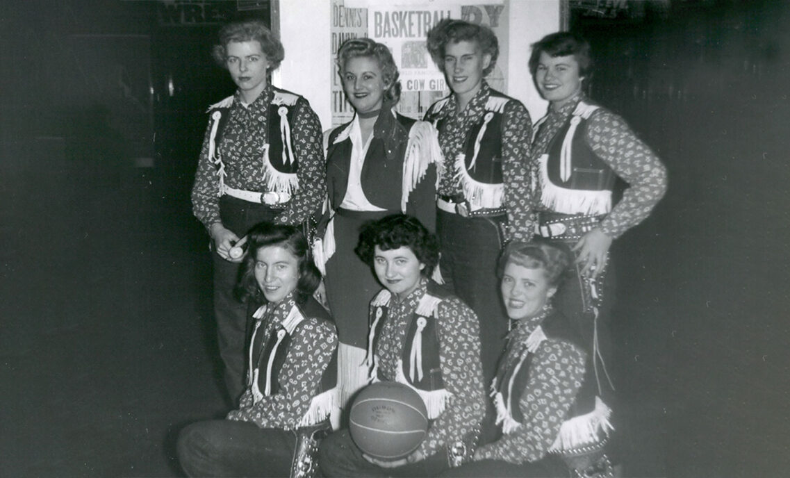 A black and white image of seven Texas Cowgirls basketball players, four standing, three kneeling in the front. The woman in the front center holds a basketball.